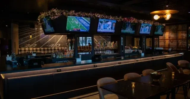 Dimly lit sports bar with illuminated bar, TVs showing football, and themed wall.