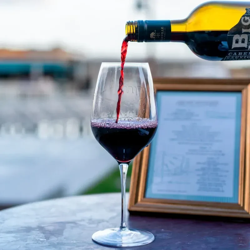 Red wine pouring from bottle into stemmed glass on outdoor table.