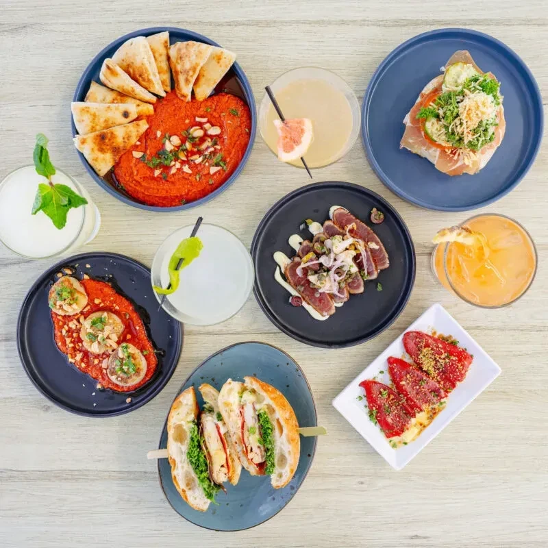Overhead flat-lay of diverse food and cocktails on a wooden table.