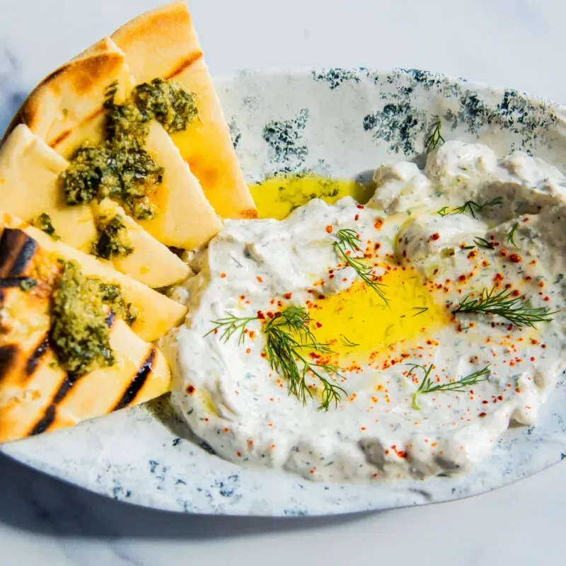 Overhead: creamy dip, dill, pita with green pesto on decorative plate.