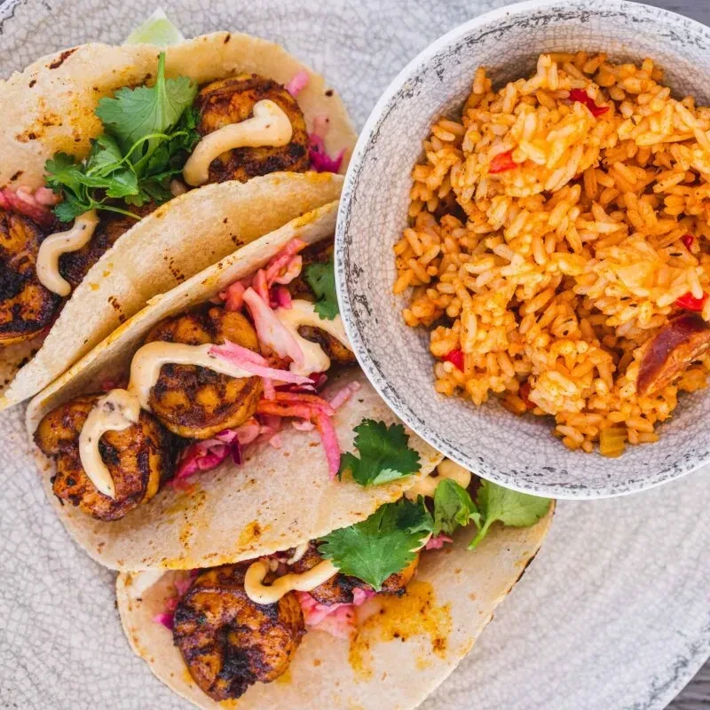 Three shrimp tacos with charred shrimp, colorful toppings, and orange rice.