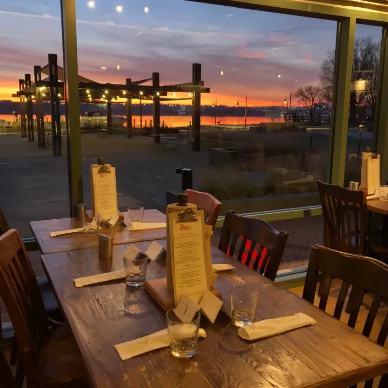 Empty restaurant tables overlook a colorful sunset over water.