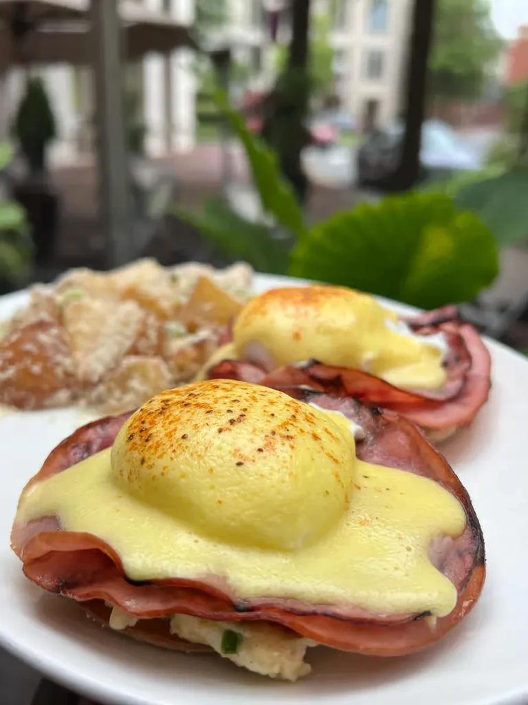 Two servings of Eggs Benedict with ham, hollandaise, and blurred outdoor background.