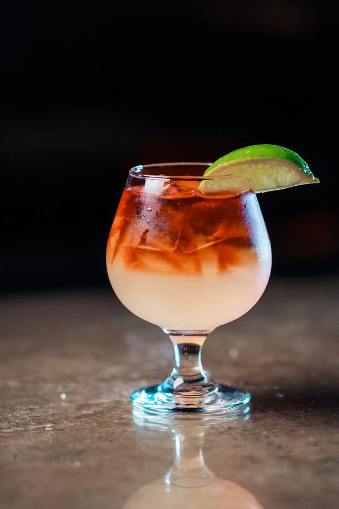 A layered white and amber cocktail with ice and a lime wedge.