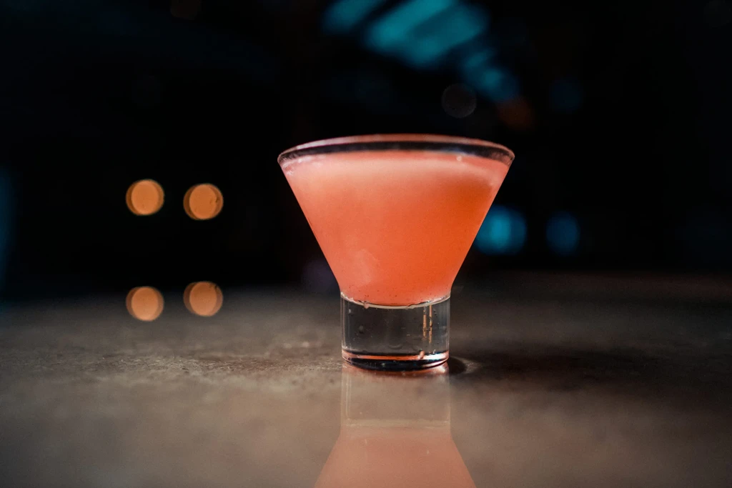 Pinkish-orange cocktail in martini glass on reflective surface, blurred background.
