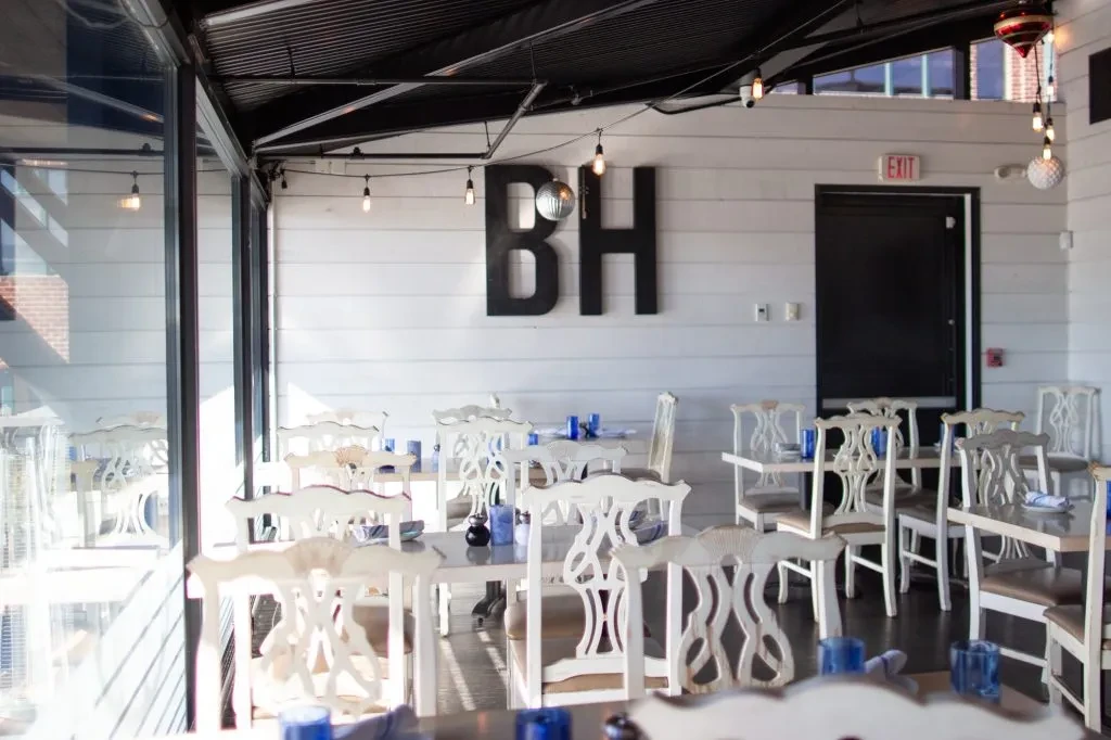 Empty bright restaurant with ornate white chairs, industrial ceiling, and 'BH' sign.