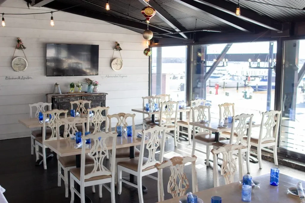 Bright, empty coastal restaurant with marina view and distressed wooden furniture.