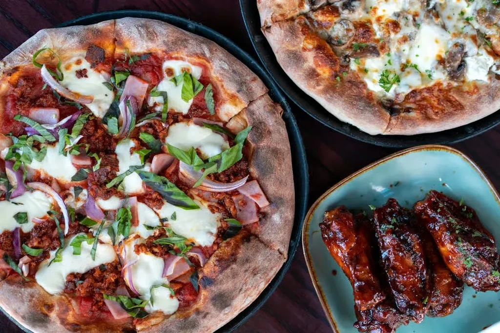 Top-down: two pizzas (red/white sauce) and glazed chicken wings.