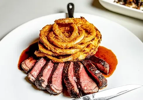 Fanned medium-rare steak with sauce and tall golden onion ring stack.