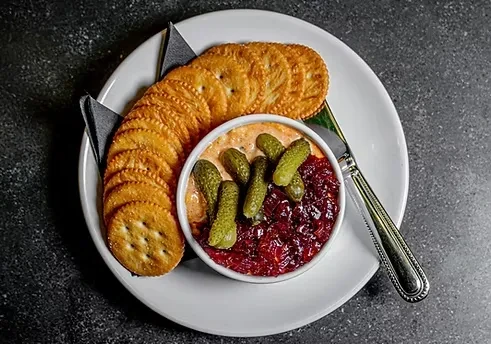Plated appetizer of savory crackers, dip with gherkins, and a knife.