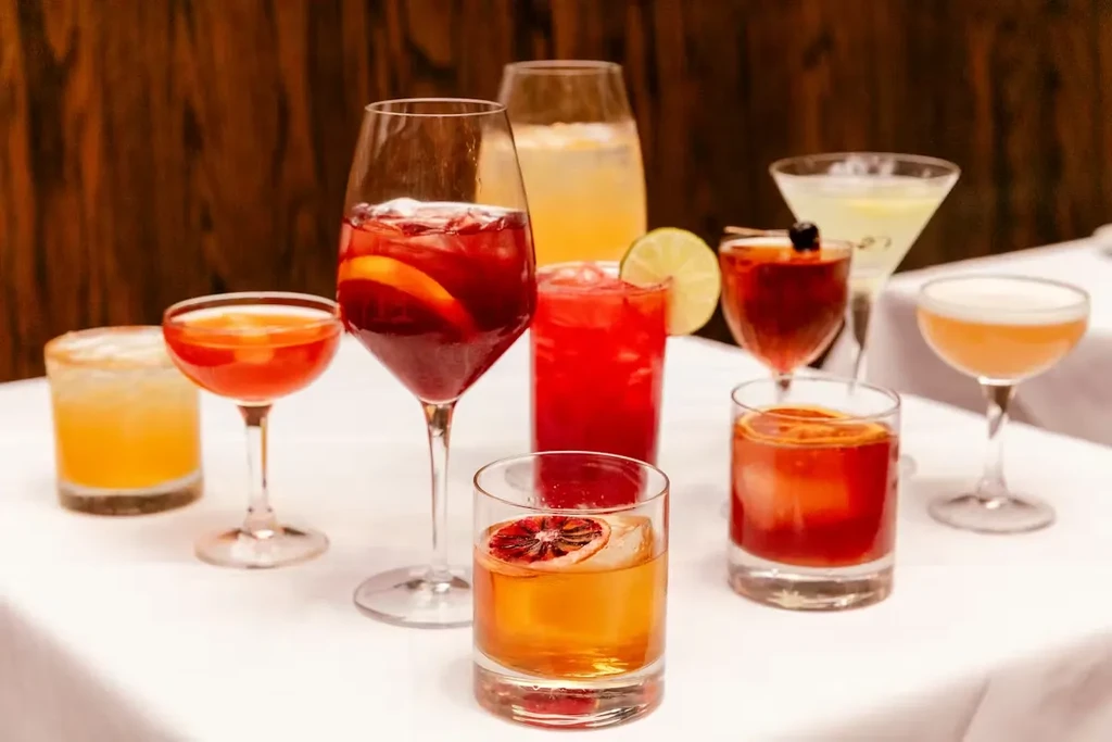 Eight colorful cocktails in various glasses, garnished, on a white tablecloth.