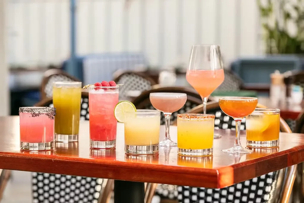 Eight colorful, garnished cocktails in various glasses on a wooden table.