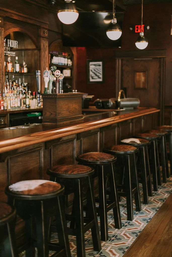 Classic bar interior with wooden counter, patterned stools, and liquor shelf.