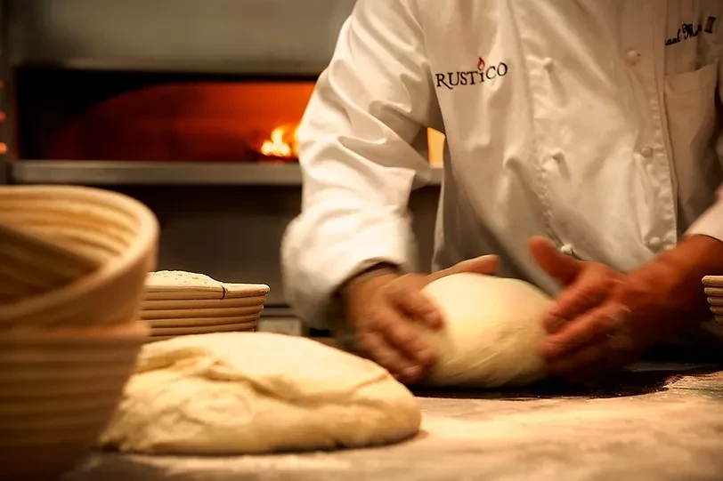 Chef's hands kneading dough on floured wood, RUSTICO logo, wood oven.