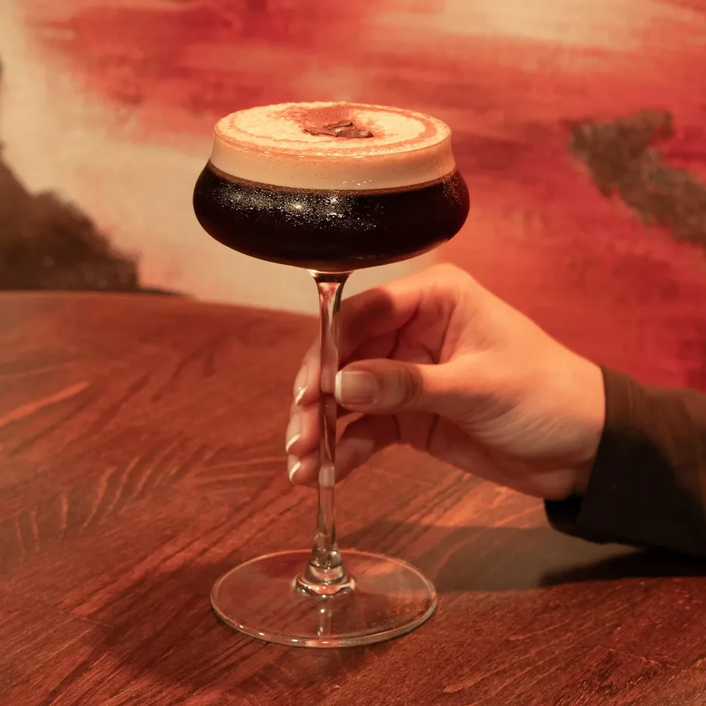Hand holds dark cocktail with foamy top in a coupe glass.