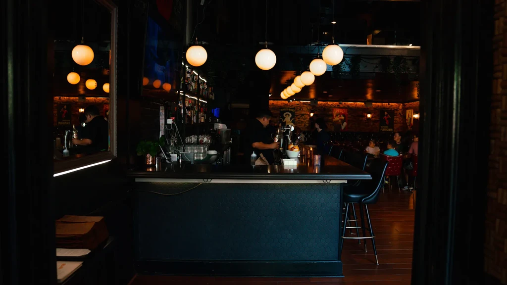 Dimly lit bar with hexagonal counter, warm globe lights, and bartender.
