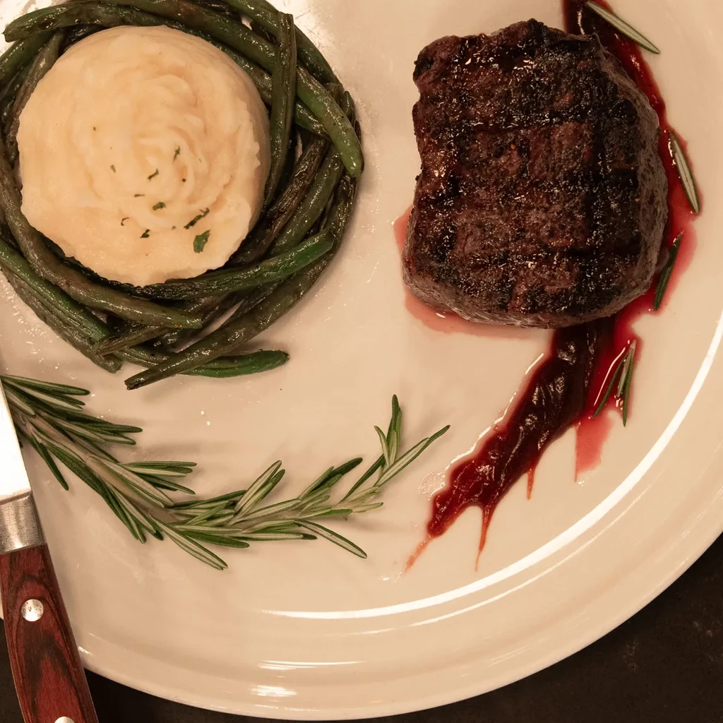 Seared steak, mashed potatoes, green beans, sauce, rosemary on a white plate.