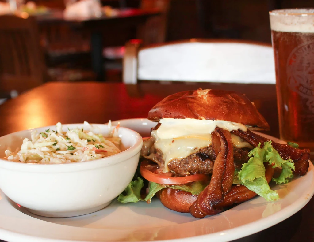 Close-up: Double-patty burger on pretzel bun, with coleslaw and beer.