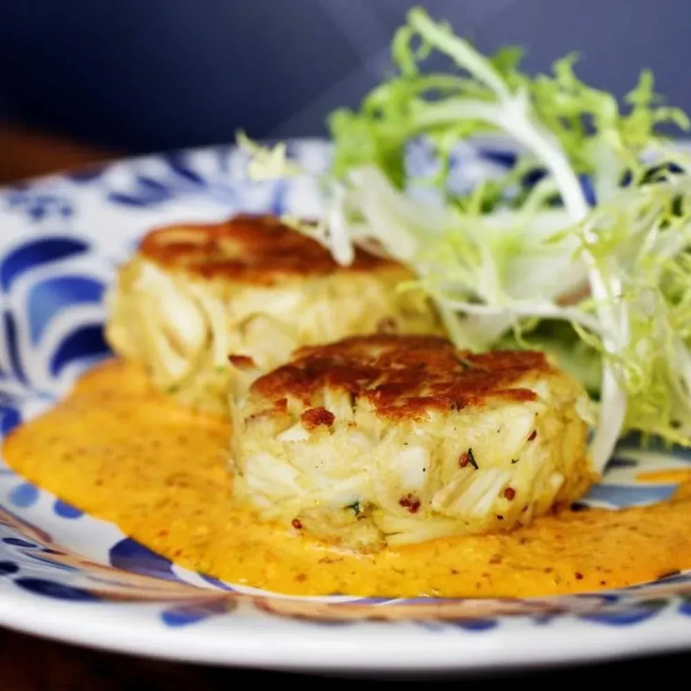 Two golden-brown crab cakes with orange sauce and frisée on a decorative plate.