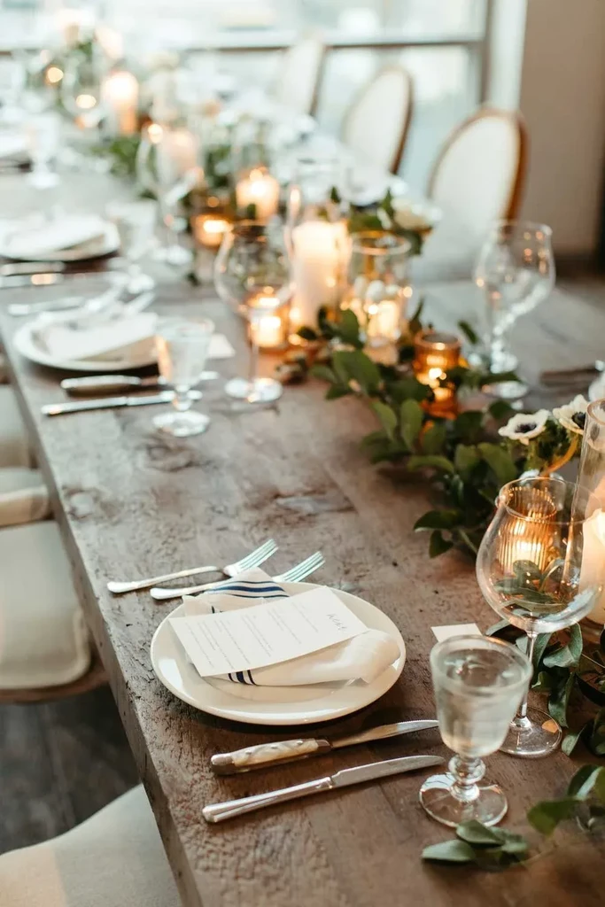 Elegantly set long wooden dining table with green garland and candles.