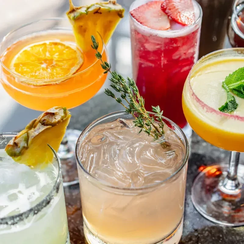 Five vibrant, beautifully garnished cocktails on a dark tabletop.