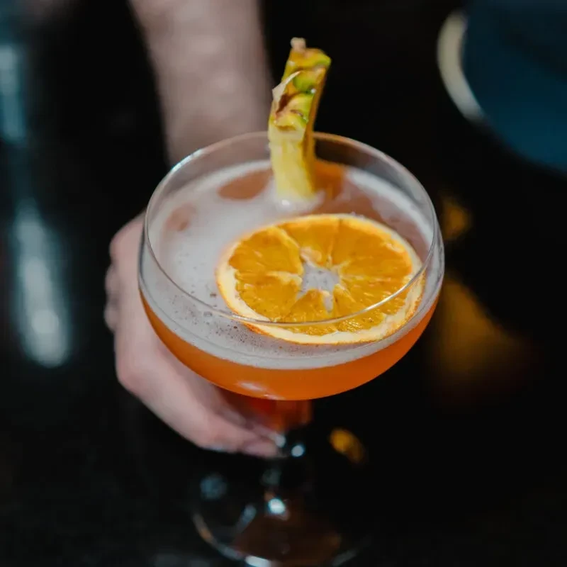 Hand holding orange cocktail, coupe glass, foam, orange, pineapple.