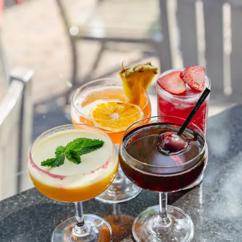 Four distinct, colorful cocktails with garnishes on a dark surface.