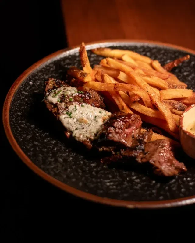 Steak pieces, french fries, and creamy herb sauce on a dark plate.