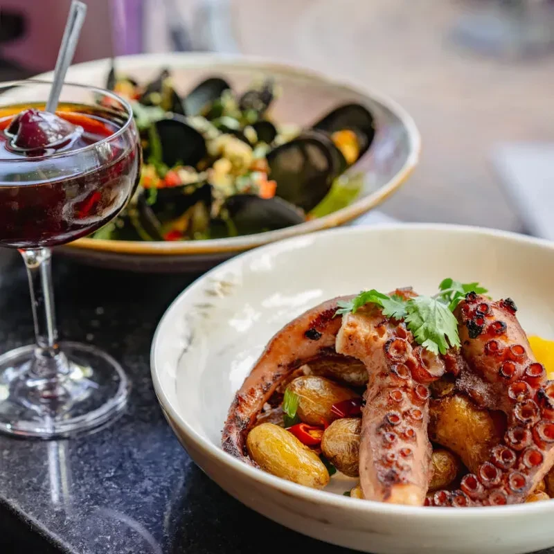 Close-up of grilled octopus, steamed mussels, and a drink on a stone table.