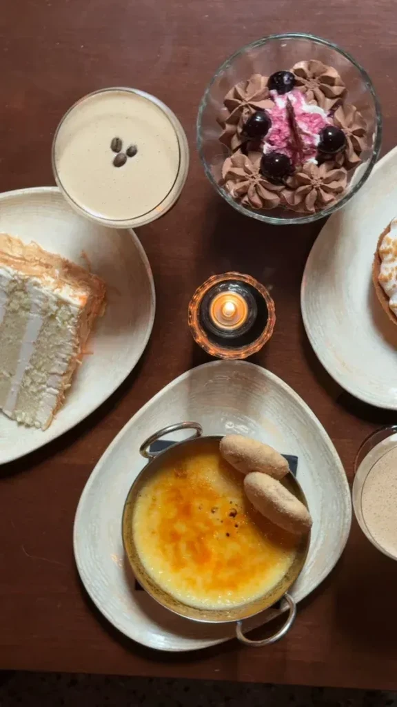 Overhead shot of a wooden table with five desserts, two creamy drinks, and a candle.