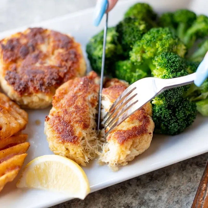 A person cuts a flaky crab cake, with broccoli and fries.