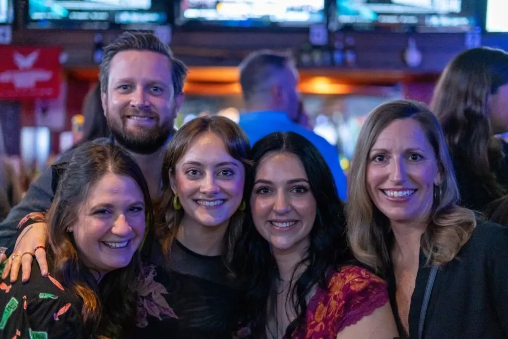 Five smiling people, one man and four women, in a dimly lit bar.