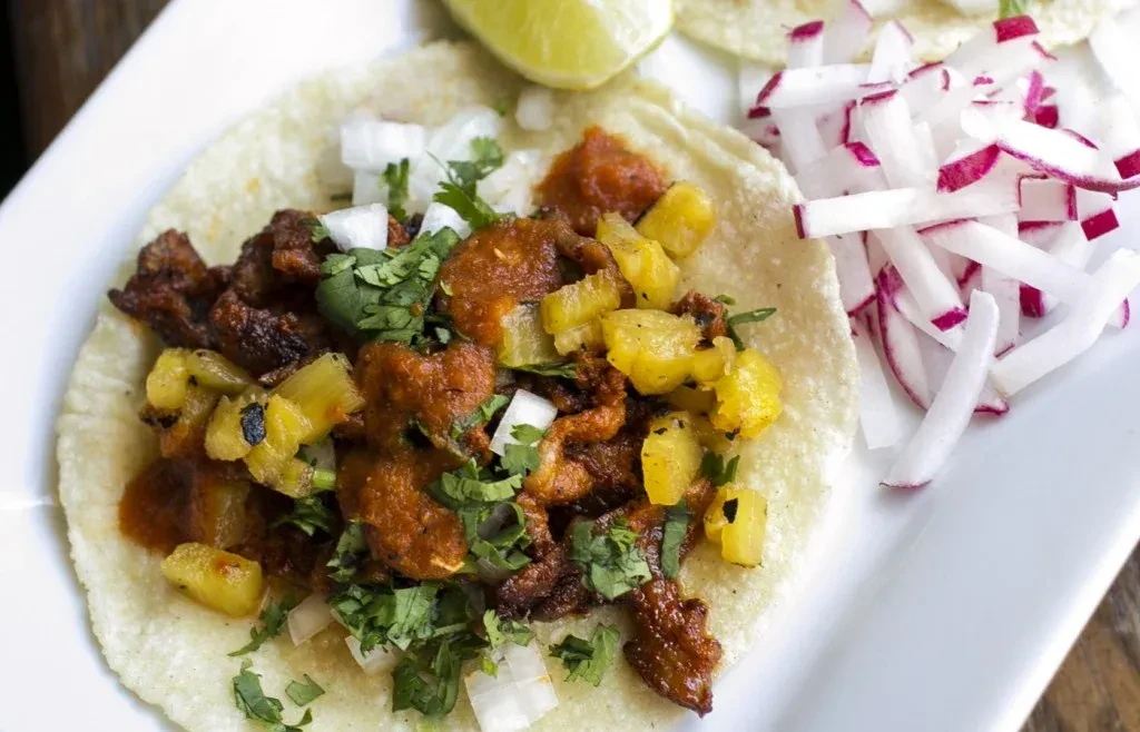 Single taco on a plate with meat, pineapple, cilantro, lime, radishes.