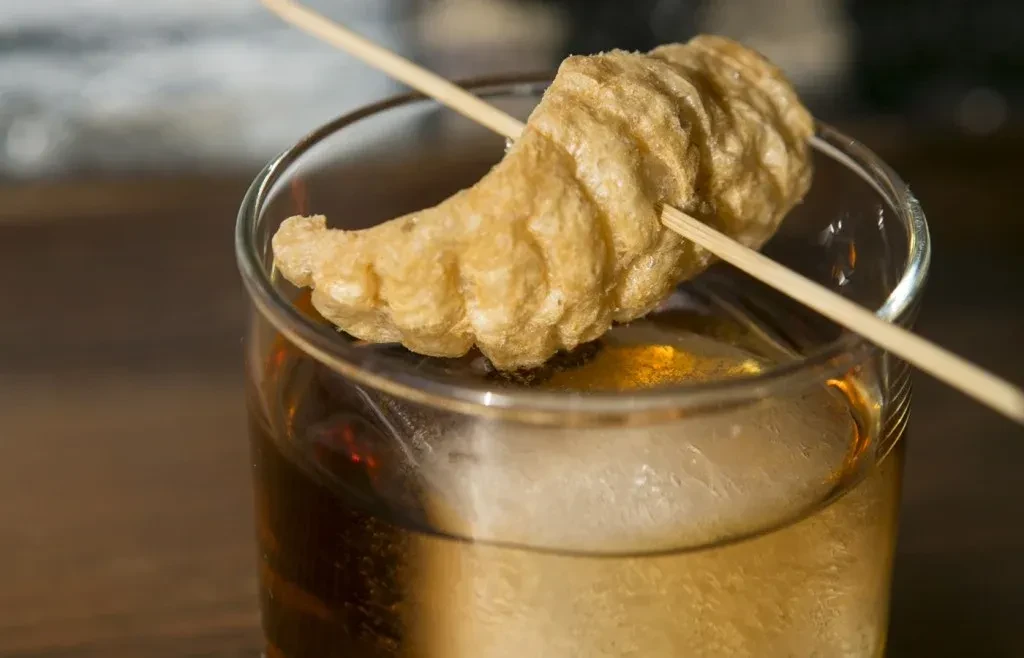 Amber drink with large ice cube, garnished with crispy chicharrón.