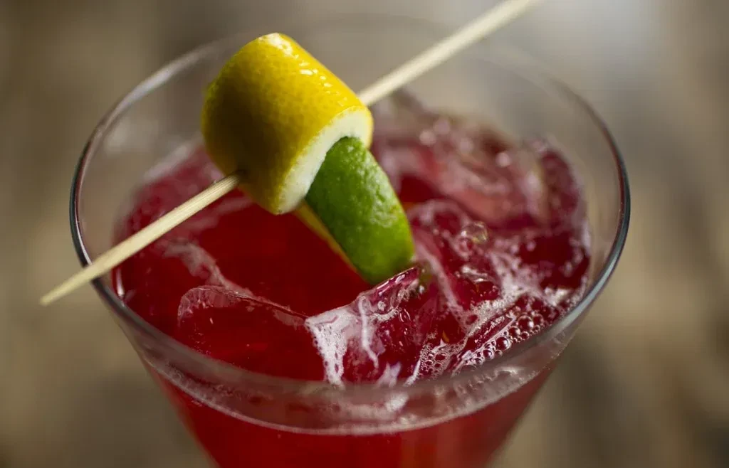 Red cocktail in martini glass with ice, lemon curl, and lime garnish.