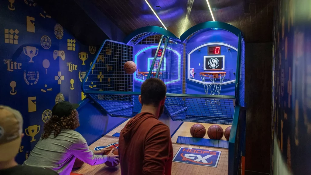 Two people play basketball arcade games in a dark, blue neon-lit room.