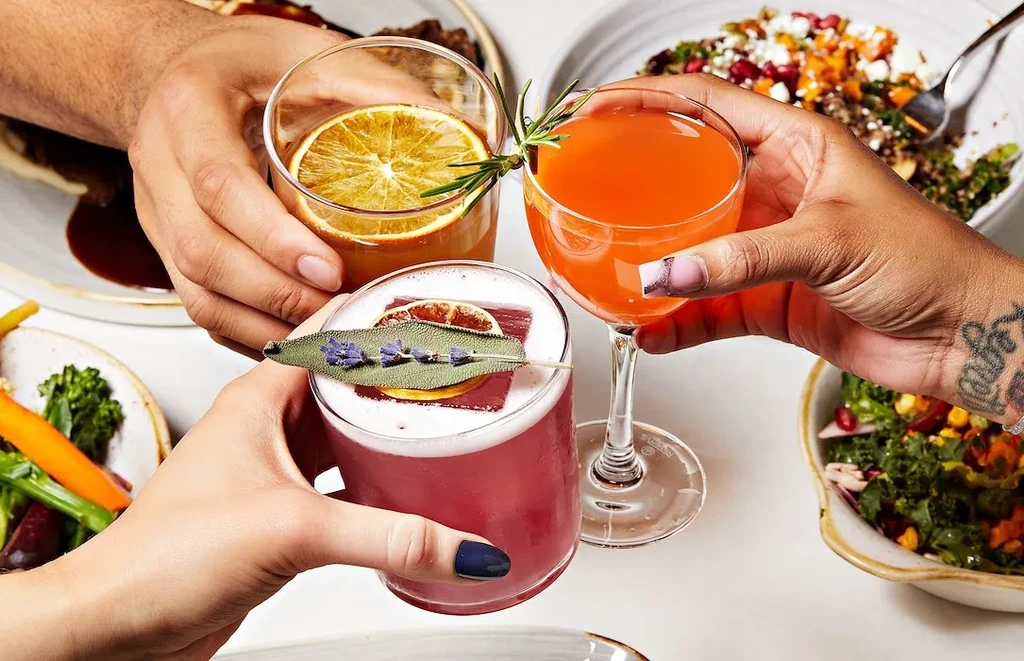 Three hands toast colorful cocktails and food dishes.