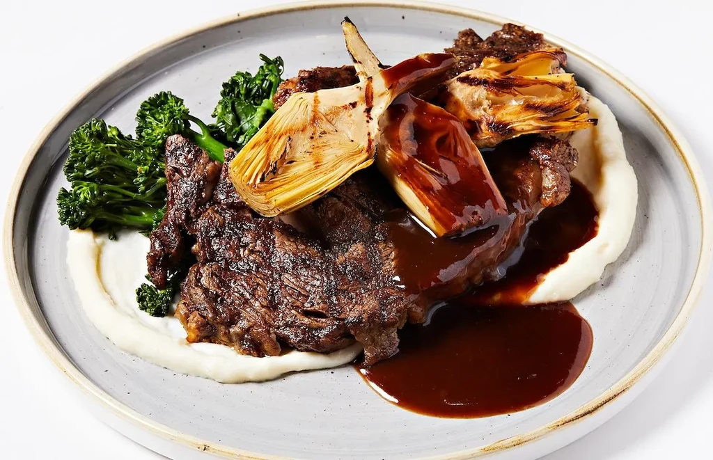 Plated braised meat, mashed potatoes, broccolini, and artichoke hearts.