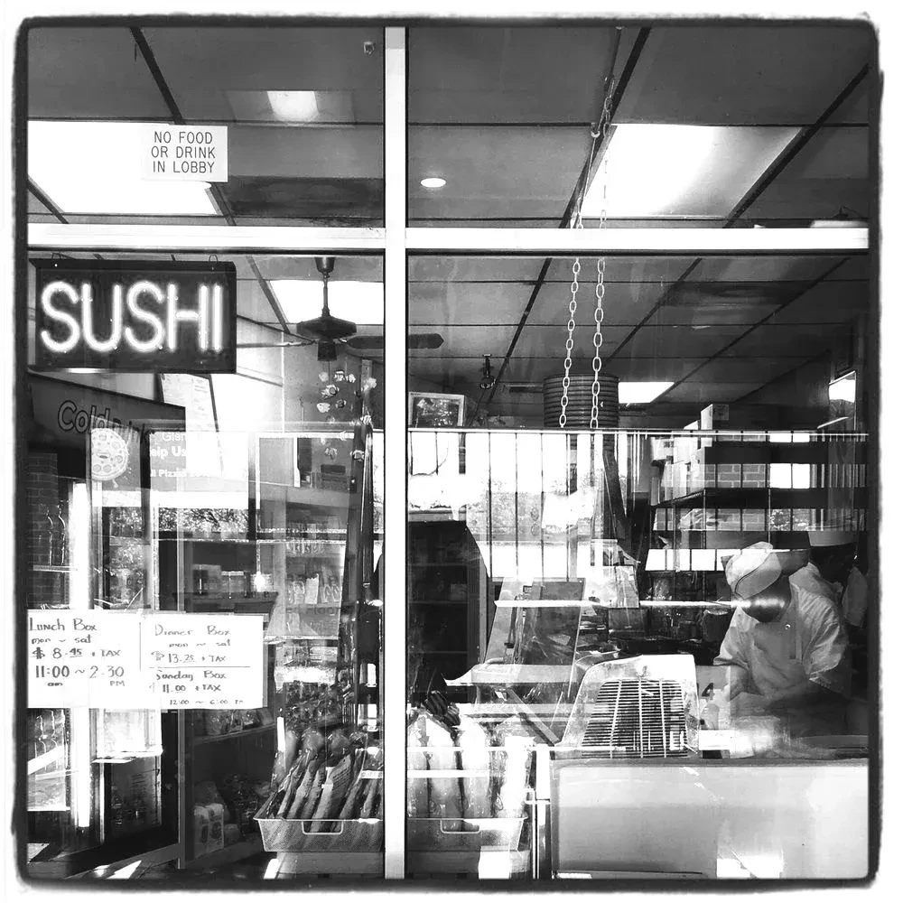Black and white photo of sushi restaurant window, signs, and chef.