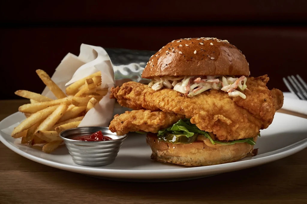 Fried chicken sandwich with coleslaw, fries, and ketchup on a wooden table.