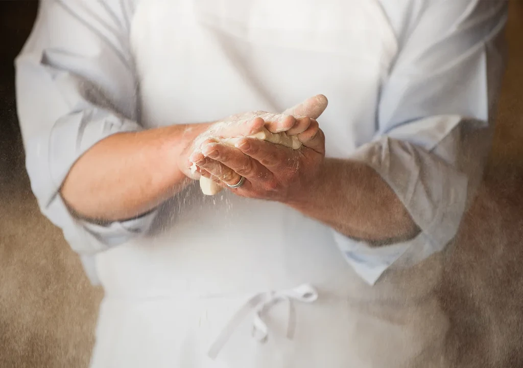 Person's flour-covered hands kneading dough, flour visibly in the air.