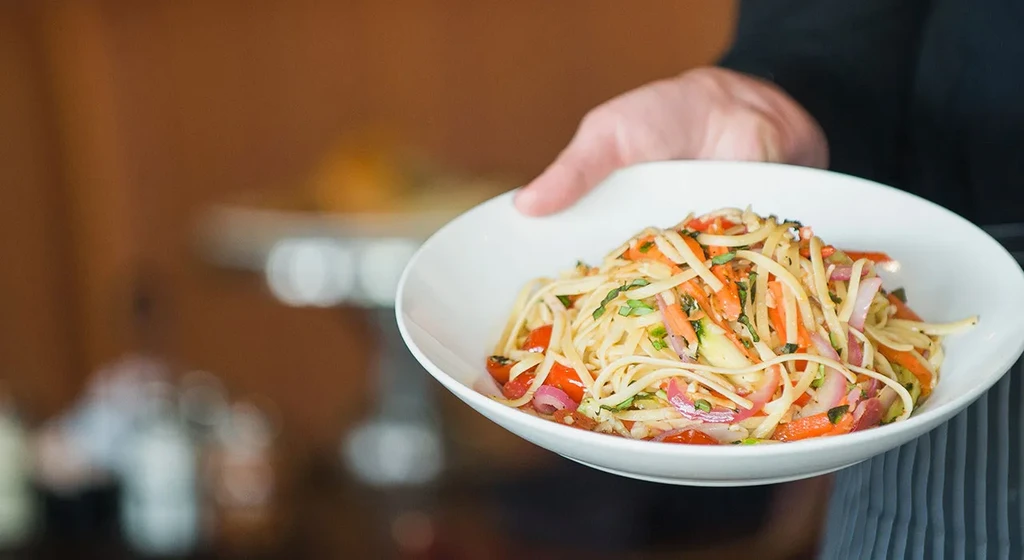 Close-up: hand holding white bowl of noodles and colorful vegetables.