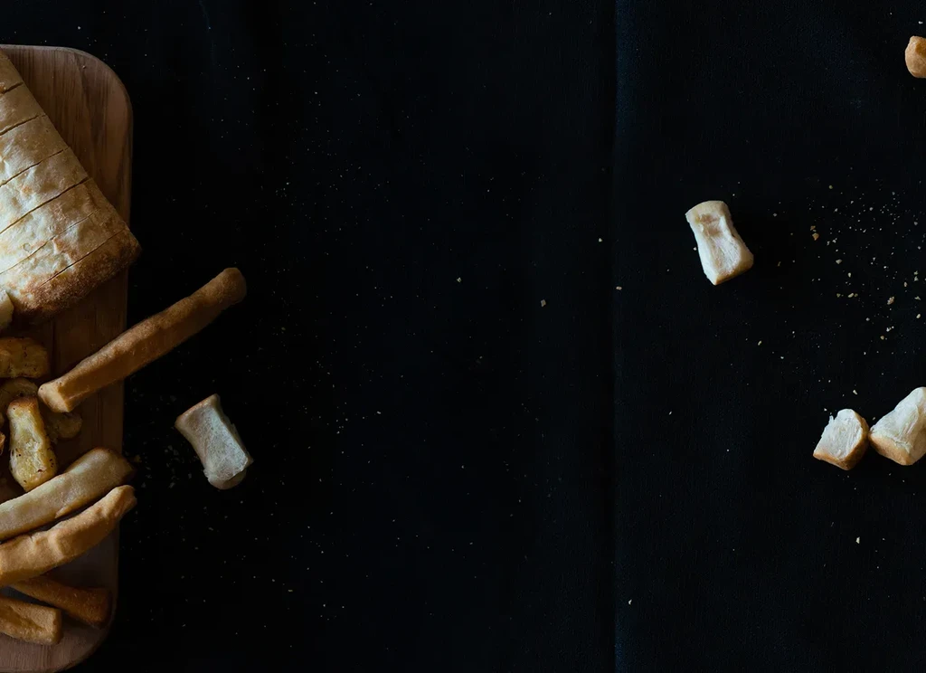Overhead, sliced bread loaf, breadsticks, and crumbs on dark surface.