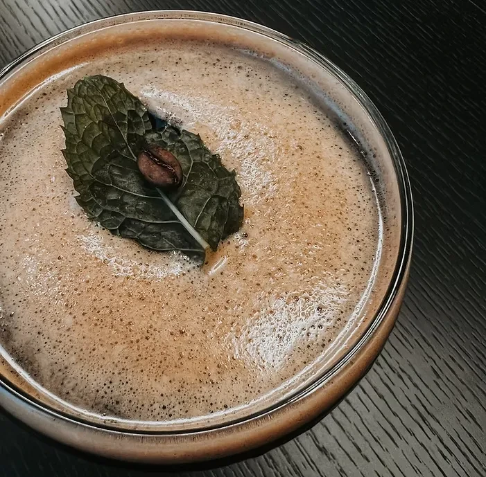 Frothy brown cocktail in clear glass, garnished with mint and coffee bean.