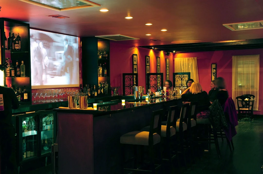 Dimly lit bar interior, magenta walls, stocked bar, projection screen, patrons.