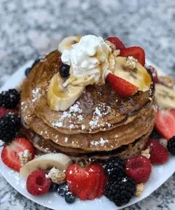Golden pancakes stacked high with berries, banana, whipped cream, powdered sugar.
