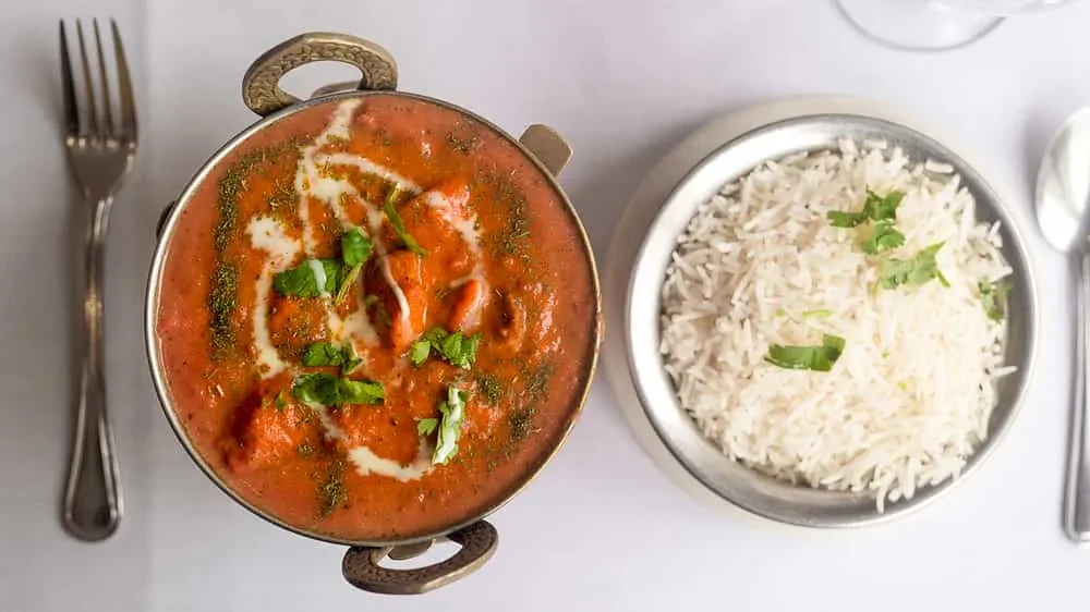 Overhead: Reddish-orange Indian curry in pot with rice, garnished on white.