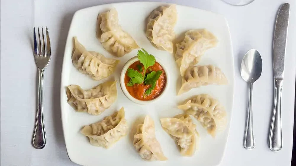 Plate of steamed dumplings, red sauce, cilantro, and silver cutlery.