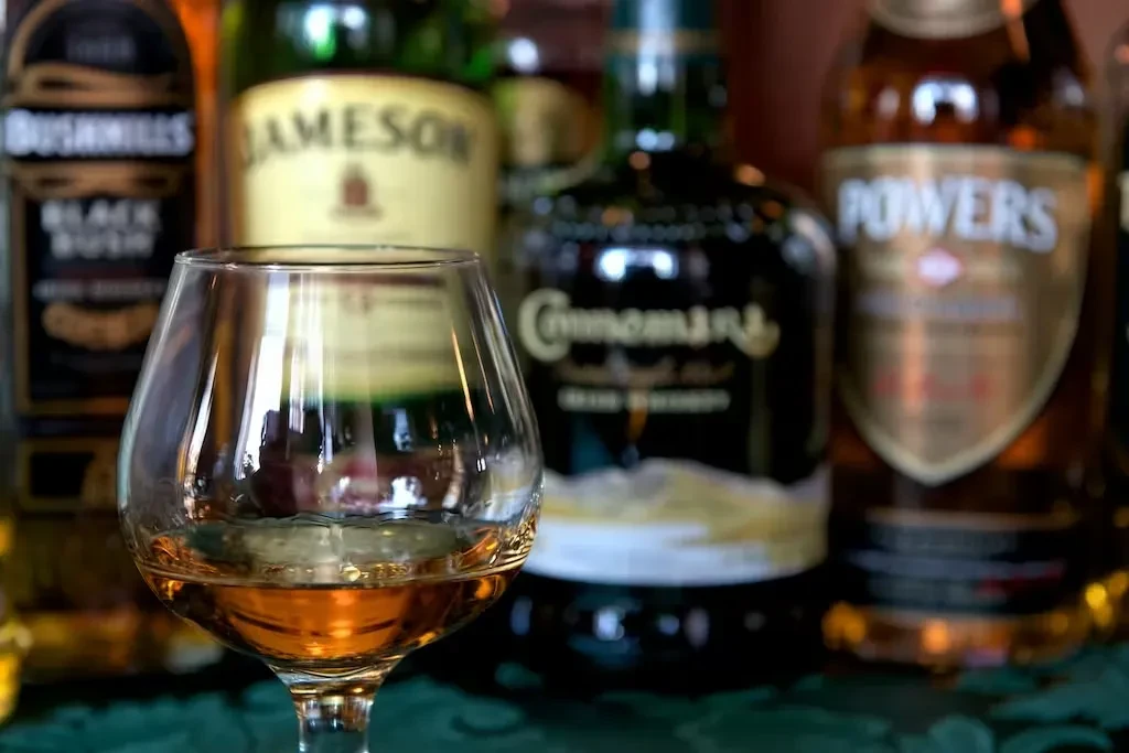 Close-up: Snifter glass of amber whiskey, blurred Irish whiskey bottles.