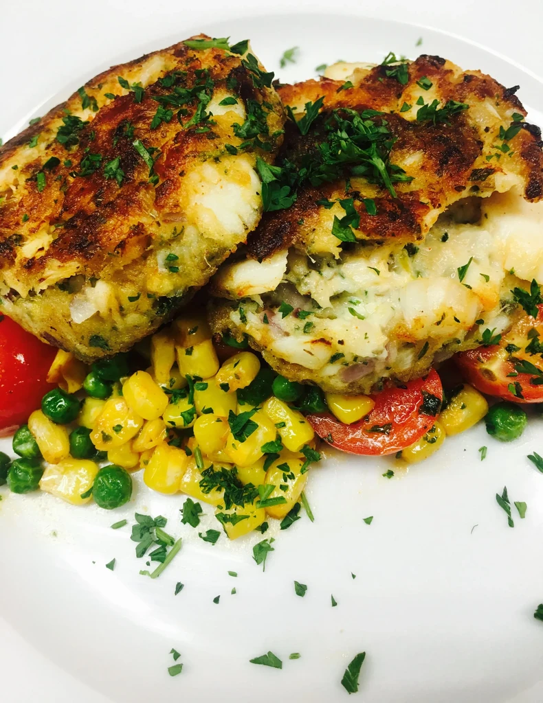 Two golden-brown crab cakes on vegetables with parsley on a white plate.